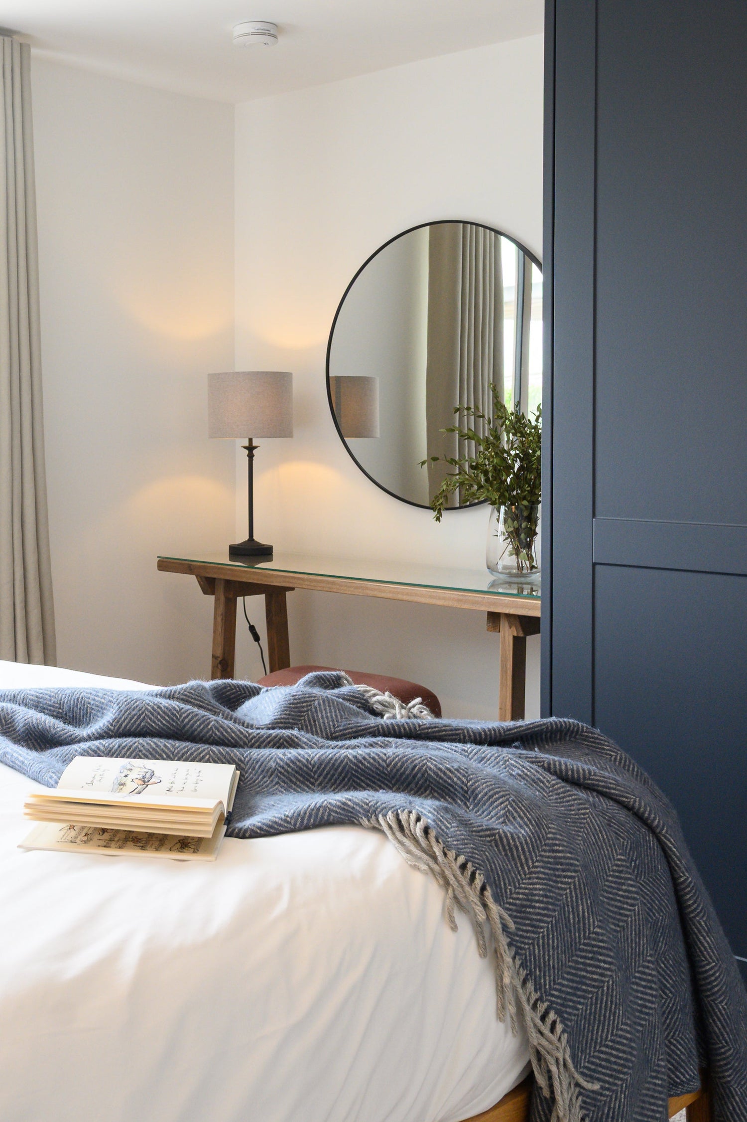 Guest bedroom with bespoke dark blue joinery, rustic wooden desk with a black frame mirror and table lamp. A dark blue wool throw covering the base of a king size bed finished in cotton sheets.